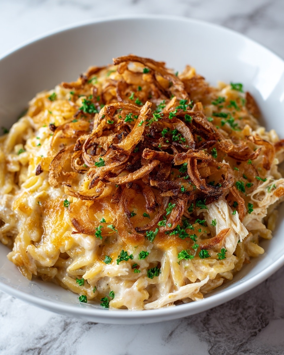 A white bowl filled with a layered pasta dish starting with a creamy mixture of short spaghetti-like noodles coated in a light beige sauce, mixed with shredded pieces of chicken. On top, there is a golden layer of melted cheese which is slightly browned, followed by a generous pile of thin, crispy fried onion rings that are dark golden and caramelized. The dish is sprinkled with small pieces of bright green chopped parsley, adding a fresh color contrast against the warm tones. The bowl sits on a white marbled surface which is visible at the edges, adding a clean and elegant look. Photo taken with an iphone --ar 4:5 --v 7