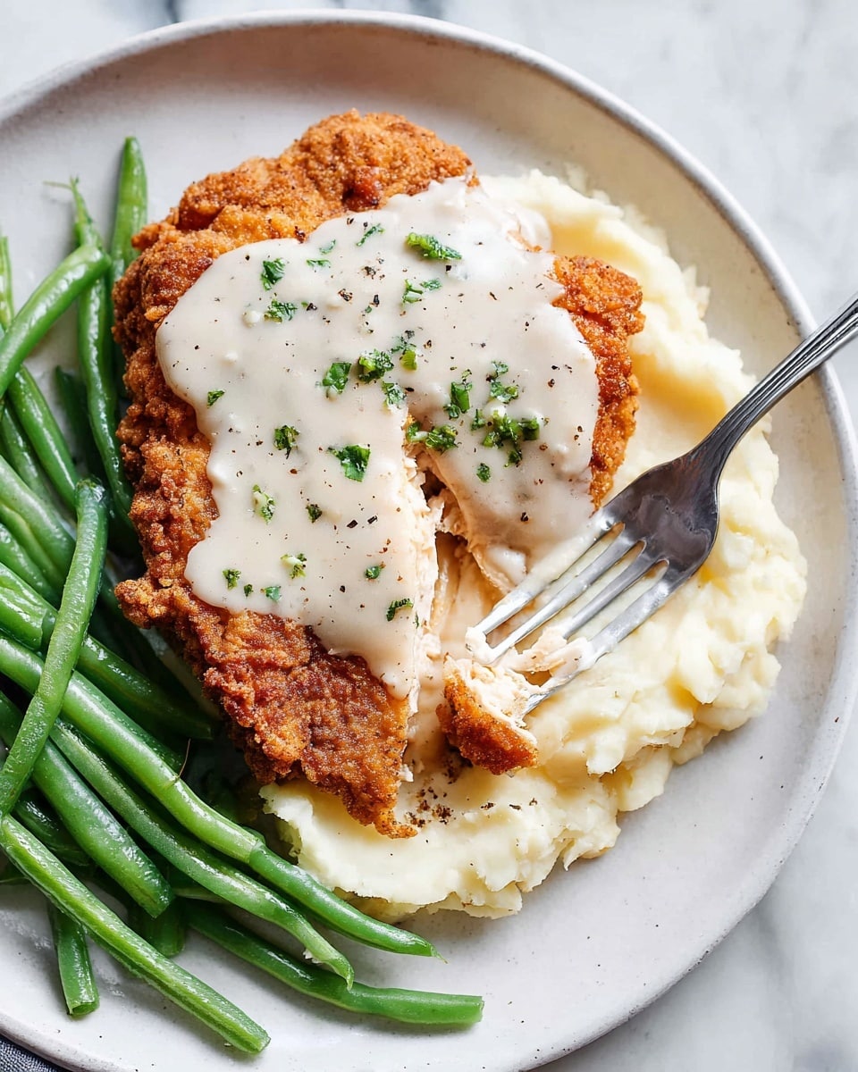 The image shows two layers of crispy golden brown fried chicken on a white plate, with a thick layer of creamy white gravy with visible black pepper spots poured over the top piece. Behind the chicken, there are bright green stalks of cooked vegetables neatly arranged. The plate sits on a white marbled surface, with the edges and texture of the fried chicken clearly visible, showing its crunchy crust. Photo taken with an iphone --ar 4:5 --v 7