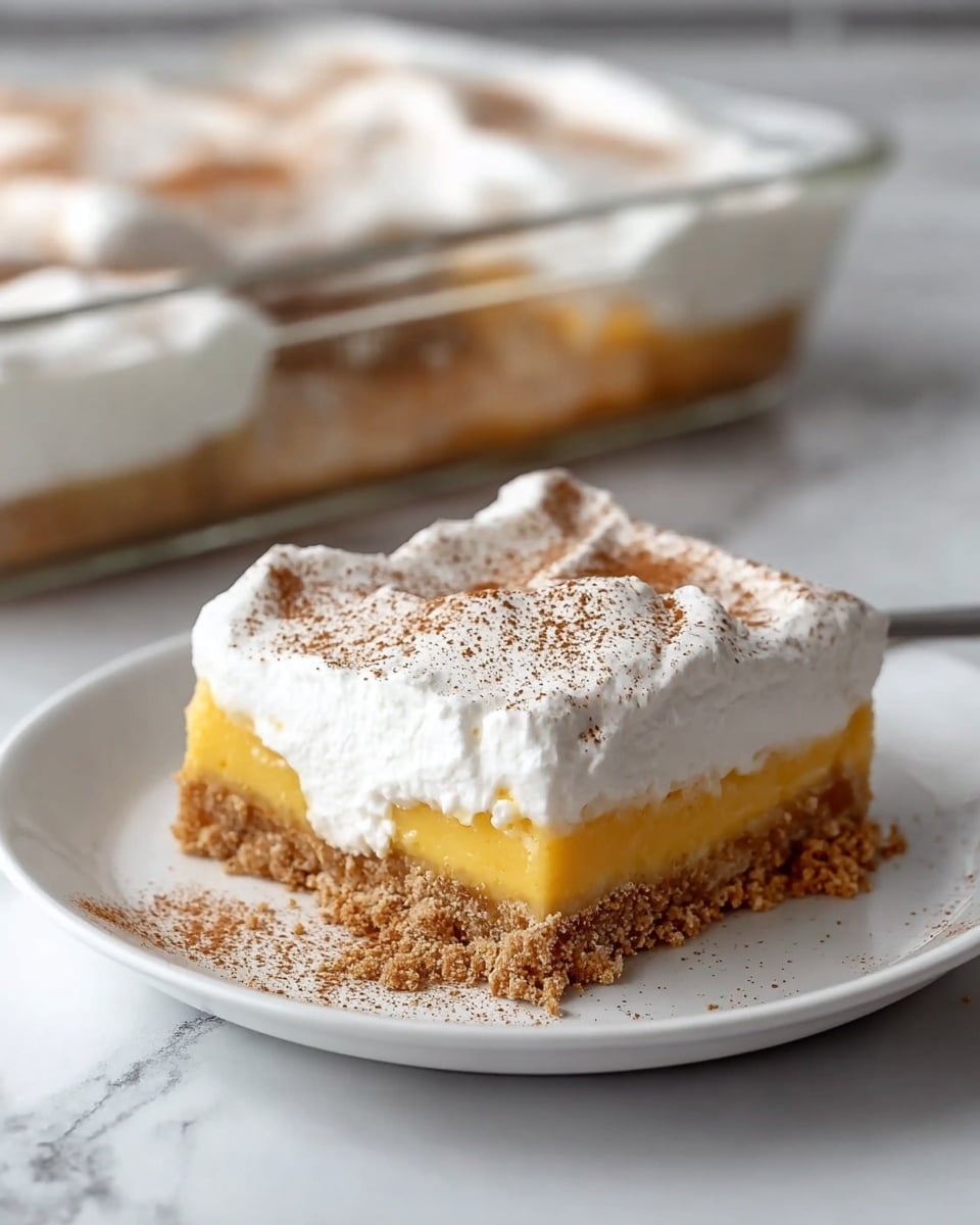 A square piece of layered dessert sits on a white plate with a white marbled surface beneath. The bottom layer is crumbly and brown, looking like a graham cracker crust. Above that is a smooth, thick yellow layer, which seems creamy and dense. The top layer is a fluffy white whipped cream sprinkled lightly with a dusting of brown cinnamon powder. The edges of the whipped cream are soft and slightly uneven, giving a fresh and airy look. In the background, a glass pan filled with the same dessert is visible, showing more of the distinct three layers. photo taken with an iphone --ar 4:5 --v 7