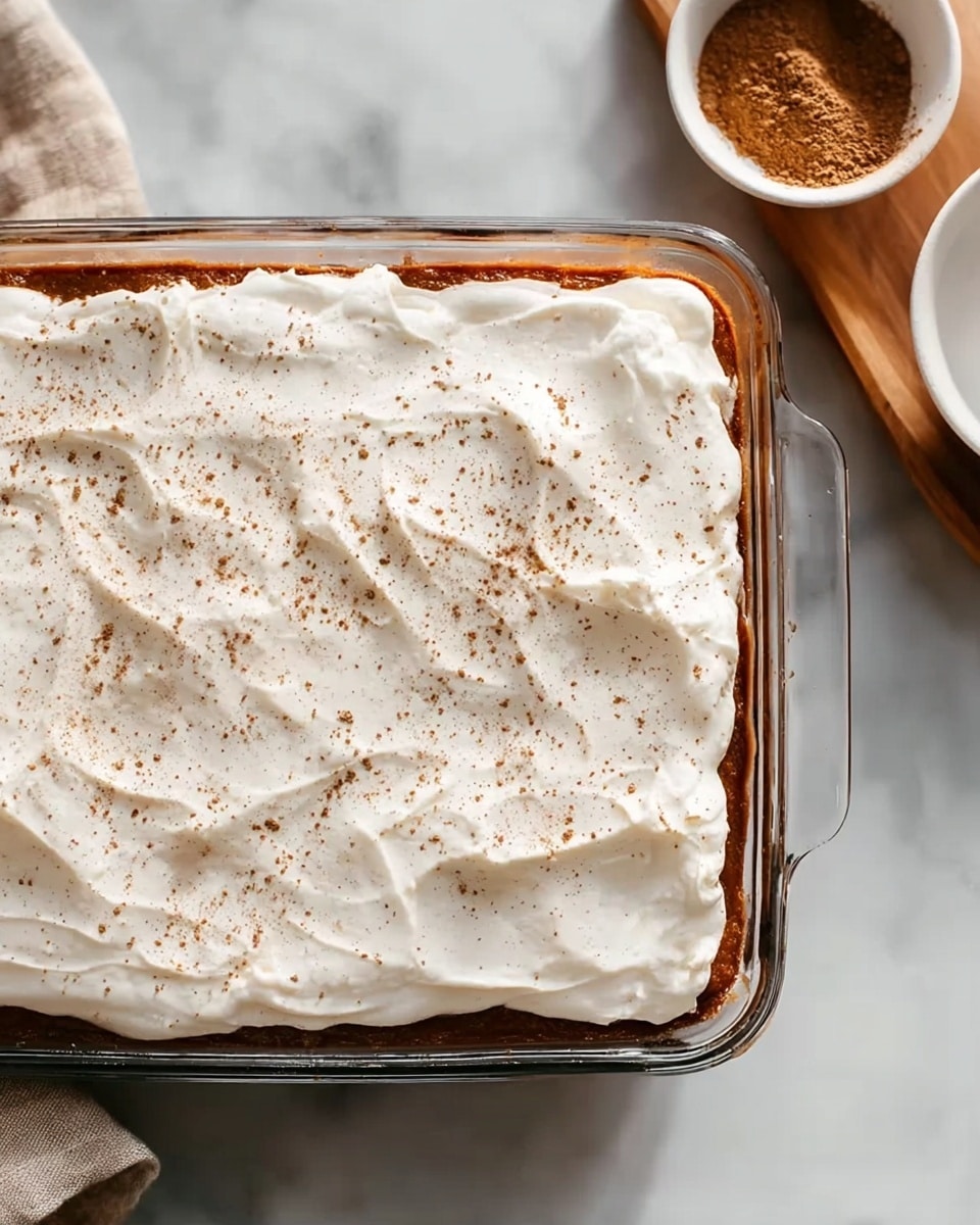 A clear glass baking dish sits on a white marbled surface, filled with a layered dessert. The bottom layer is a rich reddish-brown cake or pudding that presses slightly against the edges of the dish. On top, there is a thick, smooth layer of white cream or whipped topping, evenly spread with soft folds and small peaks. Light specks of brown spices or cocoa powder are gently sprinkled across the creamy surface, adding a touch of texture and color contrast. In the top right corner, a small white bowl holds cinnamon or cocoa powder on a wooden tray, partially visible. photo taken with an iphone --ar 4:5 --v 7
