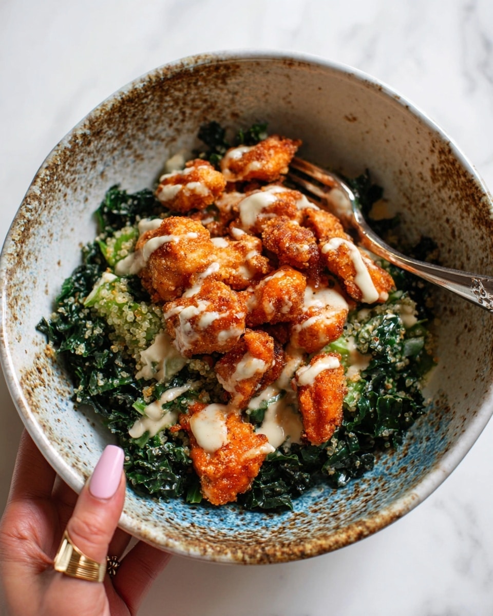 A white bowl filled with a layered dish; the bottom layer is dark green wilted kale with a slightly grainy texture, topped with small bright orange crispy battered chicken pieces lightly drizzled with a creamy white sauce, arranged in the center. The bowl is speckled with brown and blue tones and is held by a woman's hand with pale pink nails and a thin gold ring, all placed on a white marbled surface. photo taken with an iphone --ar 4:5 --v 7