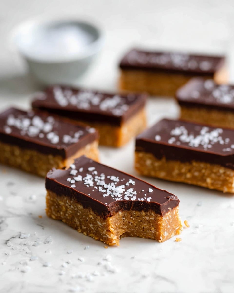 The image shows five rectangular dessert bars arranged on a white marbled surface. Each bar has two layers: a thick, rough-textured golden oat base on the bottom and a smooth, dark brown chocolate layer on top. The chocolate layer is sprinkled with coarse white sea salt. One bar in the front has a bite taken out of its corner, showing the thick layers clearly. In the blurry background, there is a small white bowl filled with coarse sea salt. photo taken with an iphone --ar 4:5 --v 7