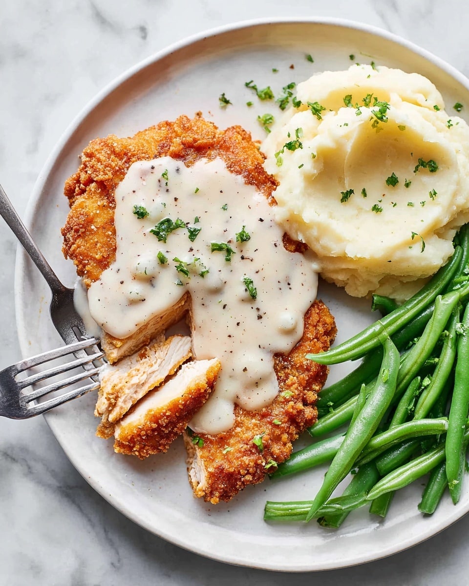 A white plate on a white marbled surface holds a three-part meal. On the left is a piece of crispy fried chicken covered with a thick layer of creamy white gravy sprinkled with black pepper and chopped green herbs, with a fork cutting into the chicken showing the tender inside. To the right is a smooth mound of creamy light beige mashed potatoes, and below it are bright green steamed green beans with a slight shine, arranged neatly. Photo taken with an iphone --ar 4:5 --v 7