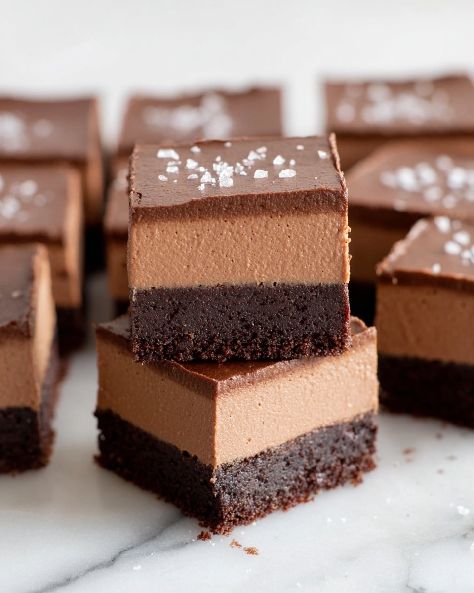 The image shows several square chocolate dessert pieces placed on a white marbled surface. Each piece has two distinct layers: a bottom dark brown, slightly crumbly layer that looks like a dense brownie or crust, and a thicker, smooth milk chocolate mousse layer on top with a creamy texture. The mousse layer has a light, creamy brown color and finishes with a slight shine, sprinkled lightly with what seems to be salt crystals. The dessert squares are neatly cut with clean edges, with three squares stacked vertically in the center while more pieces spread around in the background. photo taken with an iphone --ar 4:5 --v 7
